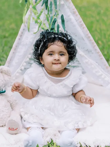 A sweet baby girl in a white dress, sitting in front of a teepee for her first birthday shoot. A simple and beautiful portrait.