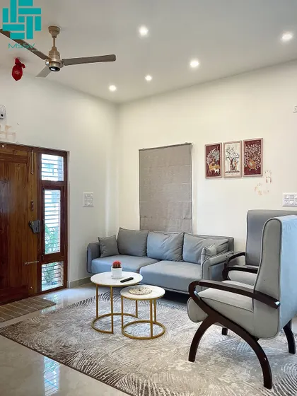 A full view of the living area, showing the traditional wooden main door and simple wall art. The design is uncluttered and functional, perfect for modern family life.