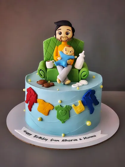 When it's the new dad's happy birthday. This vanilla and biscoff cake features a figurine of a dad relaxing on the couch with his baby, surrounded by baby clothes.
