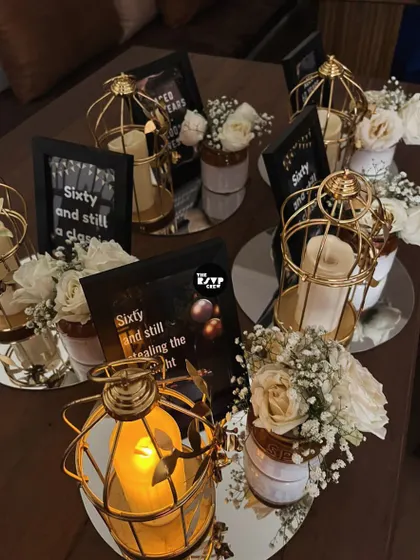Elegant table centerpieces for a 60th birthday, featuring golden birdcage lanterns, white roses, and framed signs with witty quotes like 'Sixty and still stealing the spotlight'.