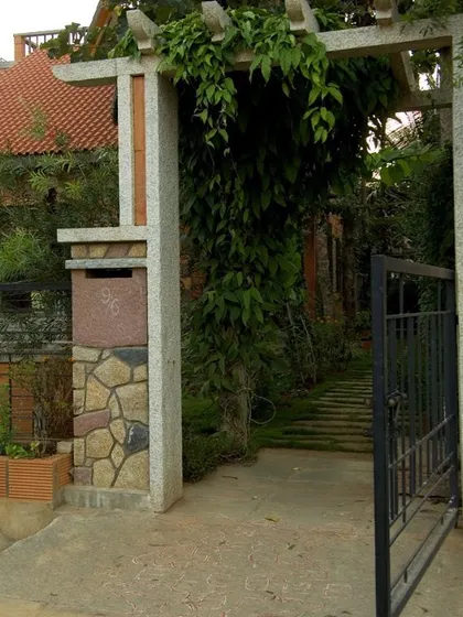The entrance gate of the Sharada Residence combines stone, concrete, and metalwork. The design includes an integrated mailbox and is topped with a pergola, which allows vines to grow and soften the structure.