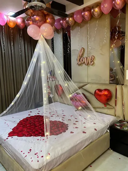 A simple yet romantic bed decoration. This setup includes a heart made of rose petals, a fairy light canopy, and foil balloons spelling 'love' on the headboard.