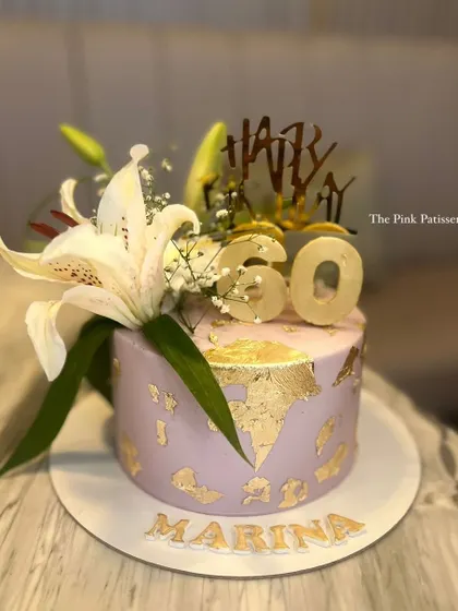 A chic 60th birthday cake in a lovely lavender shade, decorated with gold leaf, a fresh white lily, and a custom "Happy 60" topper for Marina.