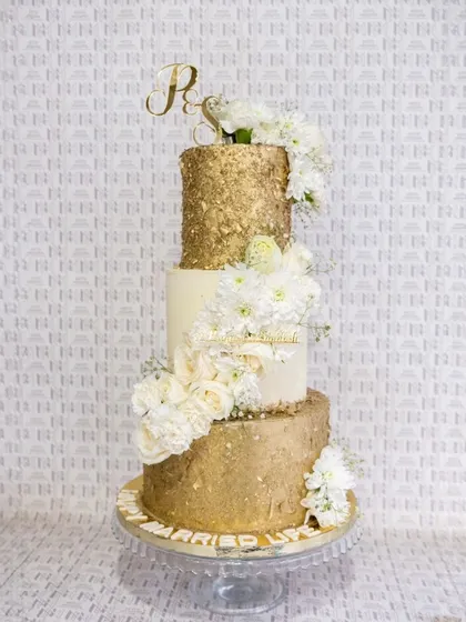 A stunning three-tiered wedding cake that combines rustic glamour with classic elegance. The top and bottom tiers are finished in a textured gold leaf, beautifully contrasted by a clean white middle tier and a cascade of fresh white flowers.