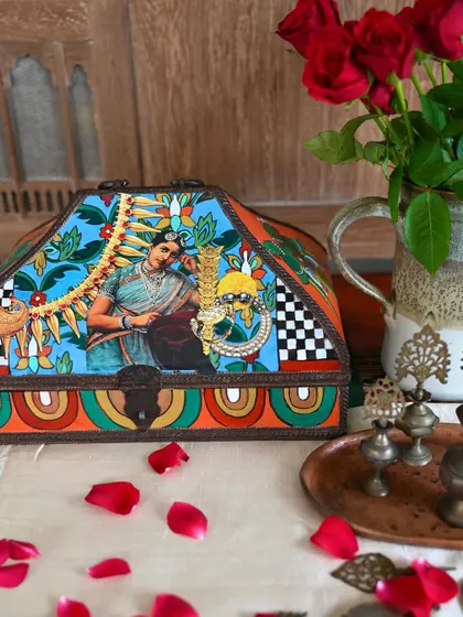 This hand-painted teak wood memory box features a portrait inspired by classical Indian art. It's a small, elegant piece perfect for a side table or dresser, shown here with fresh roses.
