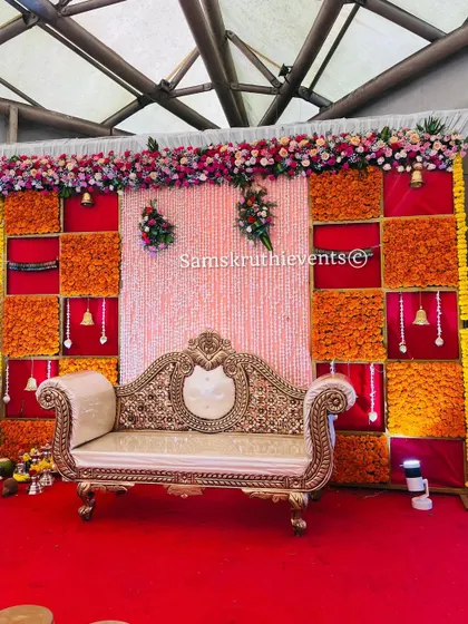 A vibrant stage decoration for a festive event. The backdrop features a checkerboard of orange and yellow marigolds against a red background, with a beautiful string of pearls curtain in the center.