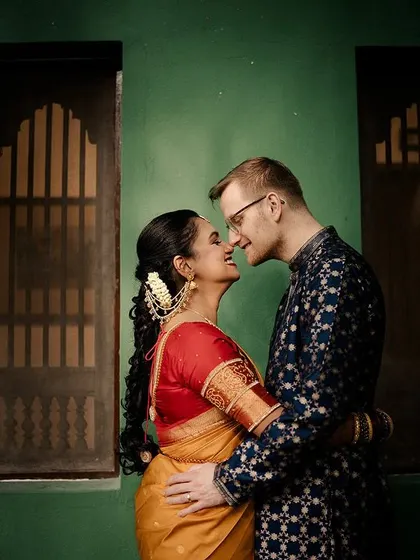 A cross-cultural couple shares an intimate moment against a green wall, their contrasting outfits telling a story of two worlds meeting.