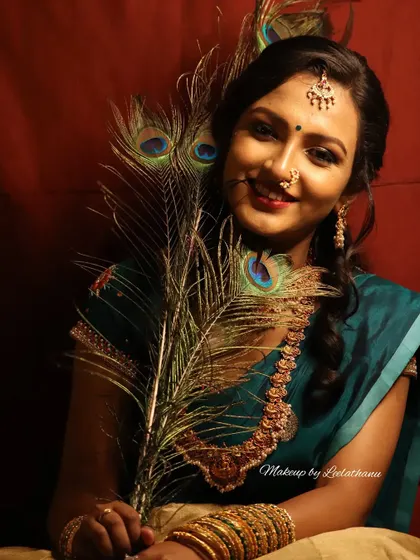 A smiling portrait from a creative shoot. The model holds a peacock feather, and her makeup has a warm, golden glow with a beautiful red bindi.