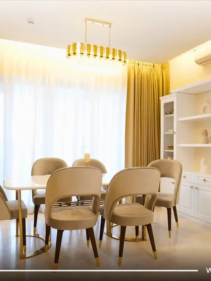 A bright and elegant dining room with cream-colored chairs, a gold-accented table, and a classic white crockery cabinet.