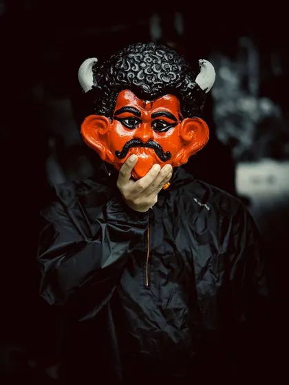 A person in a black jacket holds a traditional, brightly colored demon mask in front of their face. This moody and mysterious street portrait plays with identity and culture.