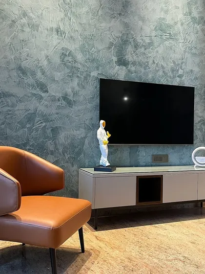 For this bedroom seating area in Ambala, we used a textured paint finish on the wall to create a dramatic backdrop. The simple TV console and tan leather accent chair provide a stylish and comfortable corner to unwind.