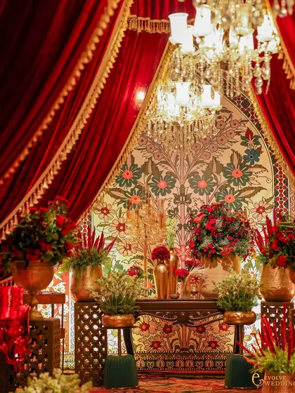An opulent entrance vignette framed by heavy red velvet curtains with gold trim. The space is filled with floral arrangements in brass vessels, all set against a beautiful tapestry backdrop.