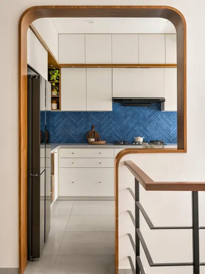 A view of the 'Palaash' kitchen through a custom curved wooden archway. The bold blue herringbone backsplash is a striking focal point, balanced by clean white cabinetry.