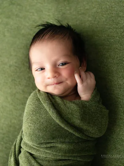 A newborn gives a sweet, sly smile while wrapped in a green swaddle. The soft, natural light beautifully highlights the curve of his smile and delicate features.