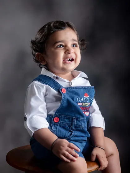 A beautiful, expressive portrait of a toddler in denim overalls. The simple, dark background and soft lighting highlight his joyful expression and bright eyes.