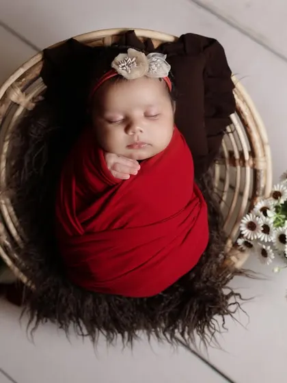 A classic newborn pose in a rustic basket. The baby is swaddled in a rich red wrap, creating a beautiful contrast with the white wooden background and floral props.