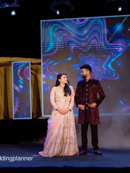 A couple on their Sangeet stage, framed by cold sparklers. The background features a large LED screen for dynamic visuals.
