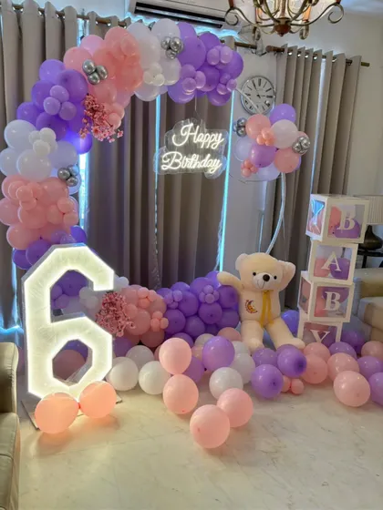 A sweet 6th birthday party setup with a pastel purple and pink theme. The decoration includes a circular balloon arch, a light-up number '6', a teddy bear, and 'BABY' blocks.