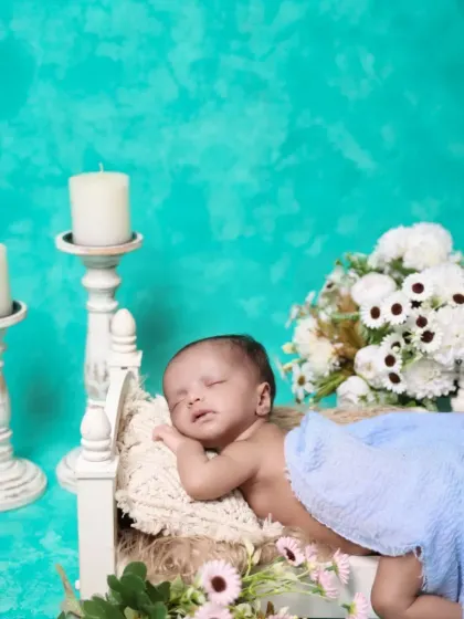 A newborn rests peacefully on a miniature bed prop against a vibrant turquoise background. The composition includes elegant candlesticks and flowers for a classic yet colorful portrait.