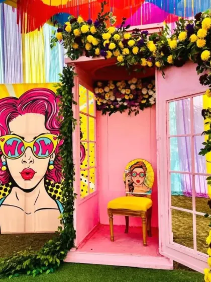 This fun photo booth was designed for a Mehandi party. It features a pink telephone booth structure adorned with yellow and white flowers, set against a pop art backdrop, creating a unique and interactive spot for guests.
