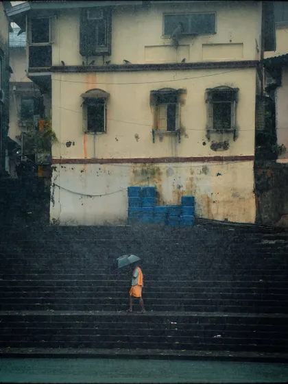 A lone figure in orange walks up the historic steps of Banganga Tank in the pouring rain. The contrast of the bright color against the muted, wet stone creates a striking visual.
