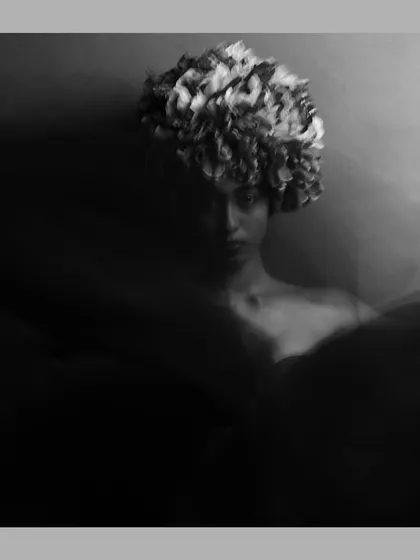 An artistic, moody portrait captured with a slow shutter speed to create a sense of motion. The model wears a floral headpiece, and the black and white treatment emphasizes the ethereal, dreamlike quality of the image.