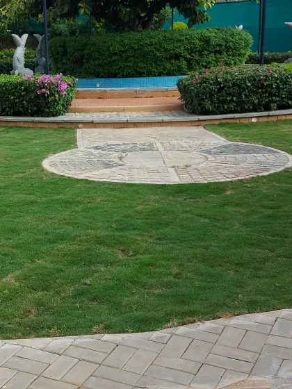 A formal garden area in a public space, featuring a circular paved design, a central water feature, and dolphin statues. The space is framed by well-maintained hedges.