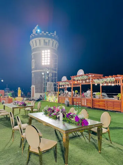 An outdoor event setup with elegant dining tables and a rustic wooden food station, set against a scenic tower.