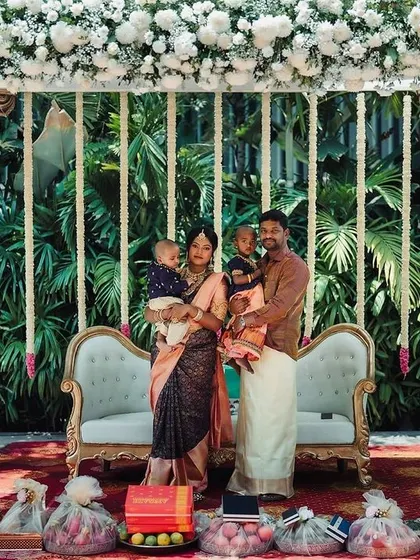A beautiful family portrait taken at a naming ceremony, featuring a traditional stage with dark wooden pillars and a canopy of white flowers.