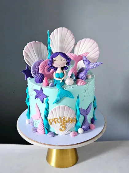 A lovely mermaid-themed cake featuring a handcrafted mermaid topper, edible seashells, and mermaid tails. The color palette of aqua, purple, and pink creates a beautiful underwater feel.