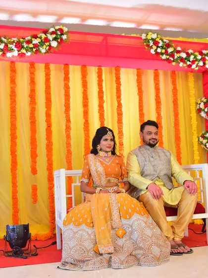 A happy couple at their Haldi ceremony. The backdrop is a vibrant mix of yellow and pink drapes with floral borders.