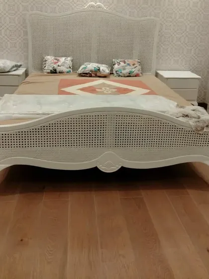 A full view of the beautiful rattan cane bed with matching side tables, set against a patterned wallpaper. The wooden flooring adds warmth to the classic white furniture.
