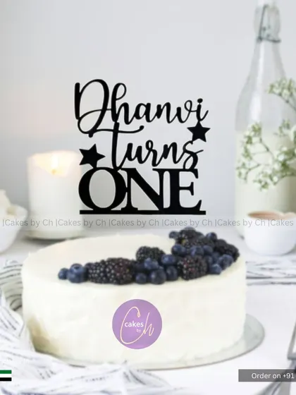 A mockup showing how the "Dhanvi turns ONE" topper in black acrylic would look on a simple white cake with berries.