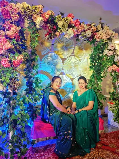 Guests enjoying a beautifully decorated photo booth. The backdrop of paper fans is framed by an arch of lush greenery and flowers, providing a lovely spot for pictures.
