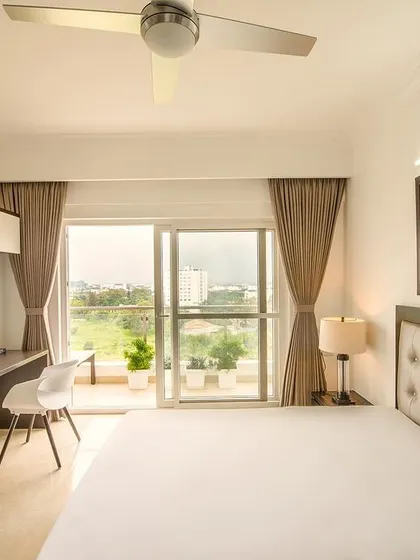 A bright and airy minimalist bedroom with a tufted headboard, a sleek integrated study desk and TV unit, and a large window offering a beautiful view.
