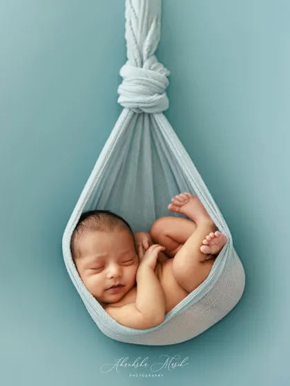 This hanging sling pose is a favorite for showing how tiny and curled up newborns are. Against a simple blue backdrop, the focus remains entirely on the baby's delicate form.