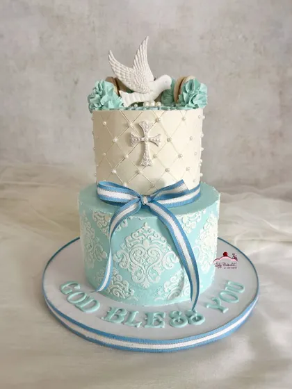 A beautiful confirmation cake in shades of blue and white. The design features intricate stenciling and a piped cross, topped with a sugar dove.