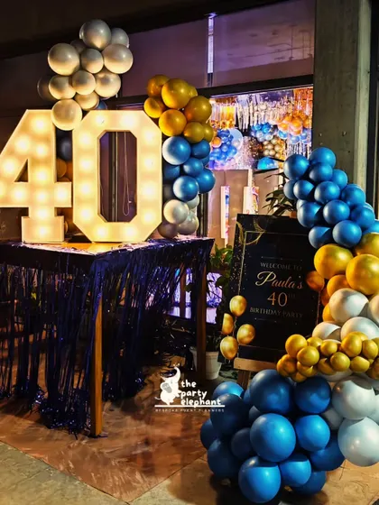 The entrance to the blue and gold 40th birthday party featured a welcome table with large marquee numbers, a personalized sign, and a cascading balloon garland to greet guests in style.