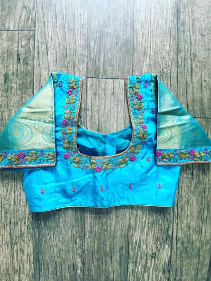 A light blue blouse with floral embroidery on the neckline. The sleeves are made from a contrasting tissue fabric with a Zari border.