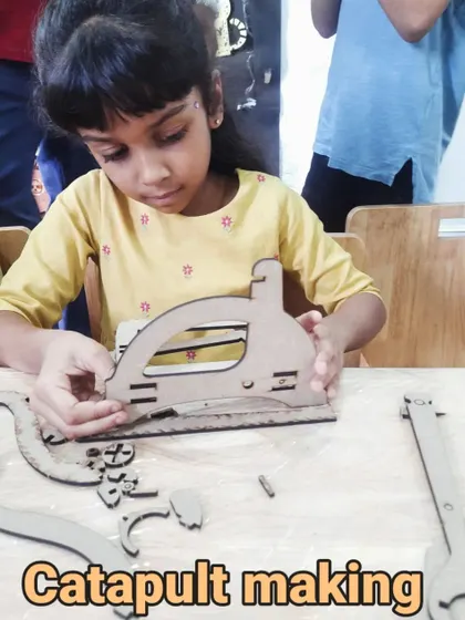 A young girl carefully assembles a wooden catapult. This project involves fitting laser-cut pieces together, teaching spatial reasoning and construction skills.