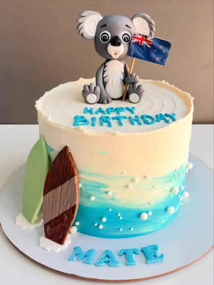 This cake takes you straight to the beach with its ocean-hued frosting and sandy details. It's topped with a cute, hand-sculpted koala holding a New Zealand flag and features two surfboards, perfect for a "Happy Birthday Mate" celebration.