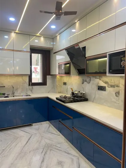 The stunning final result. A modular kitchen in a striking blue and white combination, with rose gold profile handles, a marble-patterned backsplash, and beautiful ceiling lights.