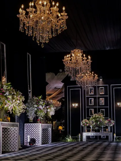 A breathtaking view of a monochrome-themed sangeet. The checkered dance floor, flanked by elegant white consoles and crowned with cascading crystal chandeliers, creates a scene of pure luxury.