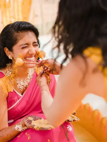 A close-up of the bride laughing as her friends apply haldi with flowers. Her genuine happiness is the highlight of this vibrant and joyful shot.