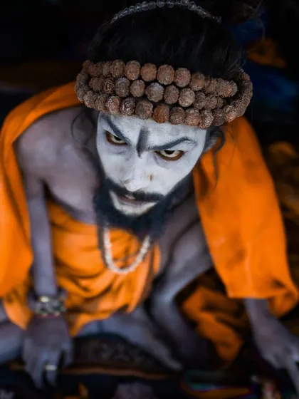 A high-angle portrait of a Naga sadhu with piercing eyes, looking up at the camera. The orange robe and ash-smeared skin create a stark and memorable image of asceticism.