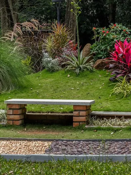 A wide view of the custom-designed reflexology pathway in a park setting. The curved path, made of different colored and textured pebbles, is set within a lush lawn and bordered by vibrant plants and a simple bench.