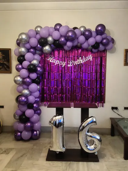 A simple and stylish setup for a 16th birthday. This L-shaped balloon arch in purple, black, and silver chrome is paired with a purple shimmer curtain and large foil number balloons.