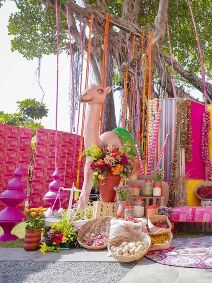 A vibrant and playful decor setup featuring a life-sized camel prop. The area is filled with colorful backdrops, hanging decorations, and baskets of spices, creating a lively market feel.