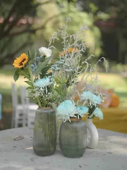 A simple yet beautiful centerpiece for a guest table, featuring sunflowers and blue-tinted flowers in green glass vases, matching the Haldi theme.