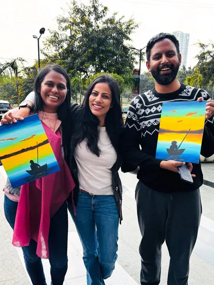 Three friends proudly showing their finished sunset paintings after a fun and creative session.